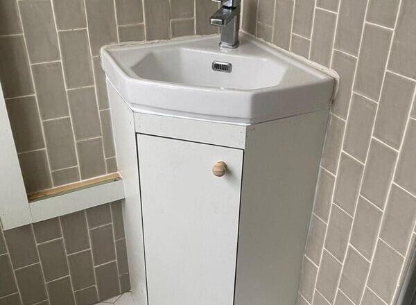 Finished white bathroom vanity cabinet fitted beneath corner sink with wooden knob.